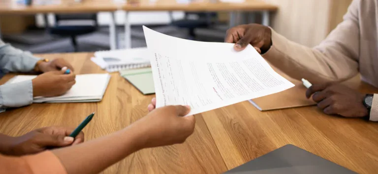 Mão de uma pessoa entregando um documento de rescisão contratual para outra pessoa sobre uma mesa de madeira.